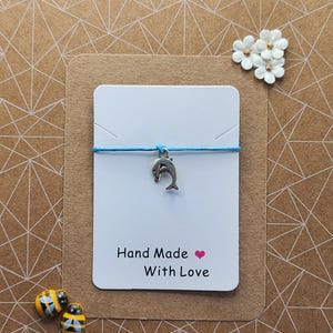 Puede incluir: Una pulsera hecha a mano con un dije de delfín plateado en un cordón azul claro. La pulsera se muestra en una tarjeta blanca con el texto "Hand Made With Love" y un corazón rosa. El fondo es una tarjeta marrón con un patrón geométrico.