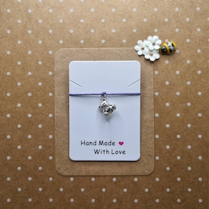 Puede incluir: Un brazalete de plata con un pequeño dije de oso. El brazalete está en una tarjeta blanca con el texto "Hand Made With Love" y un corazón rosa.