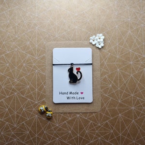 May include: A black cat charm with a red heart on a black string bracelet. The bracelet is on a white card with the text "Hand Made With Love".