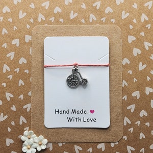 May include: A small silver charm of a tricycle on a red string with a white card that says "Hand Made with Love" and a pink heart.
