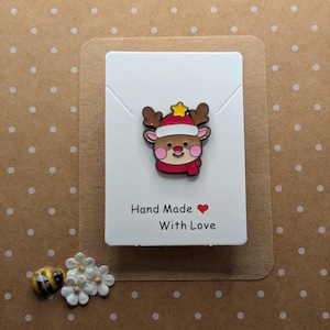 May include: A festive reindeer pin with a red hat, scarf, and a yellow star, displayed on a white card. The card has the text "Hand Made With Love" in black with a red heart. The background is brown with white polka dots.