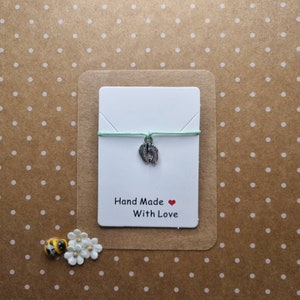May include: A white card with the text "Hand Made With Love" and a red heart. A light green string with a silver charm of two baby footprints is attached to the card.