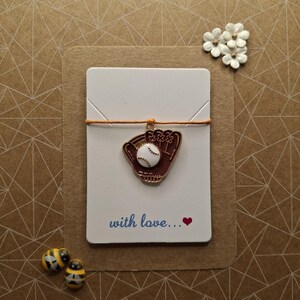 May include: A white card with a brown string tied around it. The string is holding a gold charm of a baseball glove with a white baseball inside. The card has the text "with love..." written on it in blue ink with a red heart.