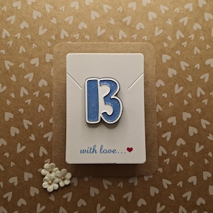 May include: A blue and white enamel pin shaped like the number 13. The pin is on a white card with the text "with love..." and a small red heart.