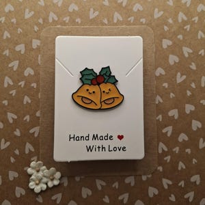 May include: A white card with a pair of yellow bells with red berries and green holly leaves. The card says "Hand Made with Love" in black text with a red heart.