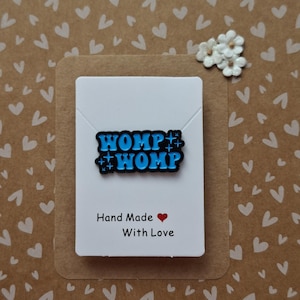 May include: A rectangular enamel pin featuring the bright blue text WOMP WOMP with small star accents on a black background. The pin is attached to a white display card that reads Hand Made with a red heart symbol and the text With Love.