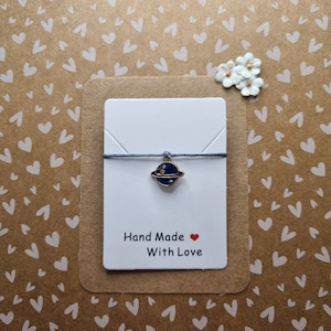 May include: A blue and gold charm of a planet with rings hangs from a grey string on a white card with the text "Hand Made With Love".