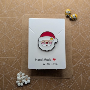Può includere: Una spilla di Babbo Natale con cappello rosso e barba bianca, su una carta con la scritta "Hand Made With Love". Sono visibili anche due spille a forma di ape e piccole decorazioni floreali bianche.