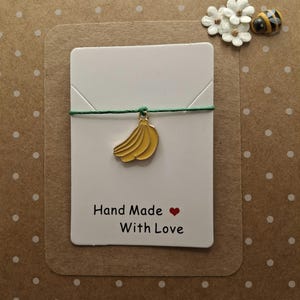 May include: A green string with a gold banana charm attached. The string is tied to a white card with the text "Hand Made With Love" and a red heart.