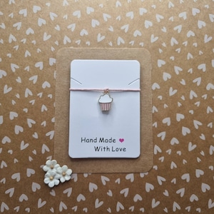 Pode incluir: Um cartão branco com um cordão rosa e um amuleto de cupcake dourado. O cartão diz "Hand Made with Love" e tem um coração rosa.