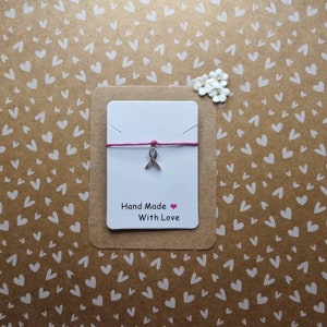 Pode incluir: Um cartão branco com um cordão rosa e um amuleto de fita prateado. O cartão diz "Hand Made With Love".