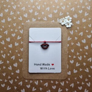 May include: A red string bracelet with a small silver charm of a red mouth with fangs. The bracelet is on a white card with the text "Hand Made With Love" and a red heart.