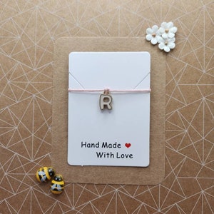 Pode incluir: Um cartão branco com a letra "R" em um cordão rosa. O cartão diz "Hand Made with Love" e tem um símbolo de coração vermelho.