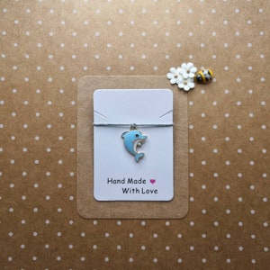 Puede incluir: Una tarjeta blanca con un dije de delfín azul en una cuerda gris. La tarjeta dice "Hand Made With Love" con un corazón rosa.