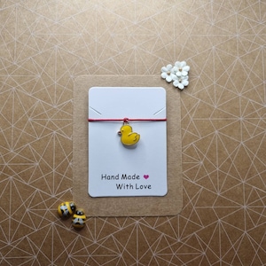 Peut inclure: Un bracelet en cordon rouge avec un charme de canard en caoutchouc jaune. La carte indique "Hand Made With Love".