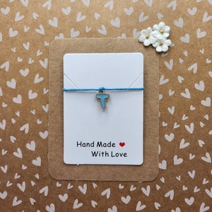 May include: A blue and gold charm bracelet with the letter "T" on it. The bracelet is on a white card with the text "Hand Made With Love" and a red heart.