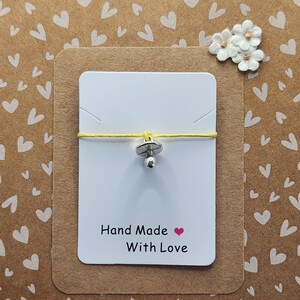 May include: A silver-toned pacifier charm on a thin yellow cord, mounted on a white rectangular display card. The card features the black printed text Hand Made with a pink heart symbol and the words With Love below it.