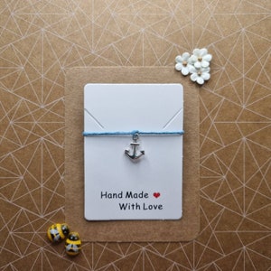 Può includere: Un biglietto bianco con uno spago blu e un ciondolo a forma di ancora argentato. Il biglietto dice "Hand Made with Love" e ha un cuore rosso.