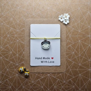 May include: A yellow string with a silver monkey charm attached. The charm has a red heart on its face. The string is on a white card with the text "Hand Made With Love".