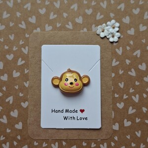 May include: A brown and tan monkey-shaped pin with pink cheeks and a small smile, attached to a white rectangular display card. The card features the black text "Hand Made" followed by a red heart icon and the words "With Love" below it.