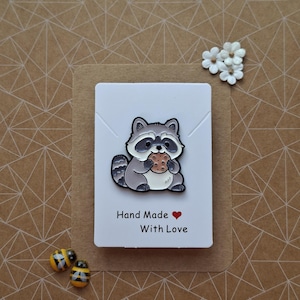 May include: A metal enamel pin featuring a cartoon raccoon with grey and white fur holding a brown chocolate chip cookie. The pin is attached to a white rectangular display card with the text Hand Made With Love and a small red heart icon.