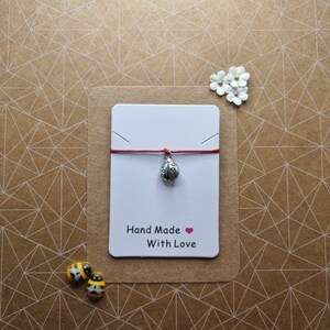 May include: A red string bracelet with a silver ladybug charm. The bracelet is displayed on a white card with the text "Hand Made With Love".