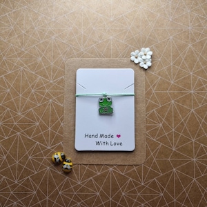 Peut inclure: Un charme de grenouille verte avec un cœur rose sur une ficelle vert clair. Le charme est sur une carte blanche avec le texte "Hand Made With Love".