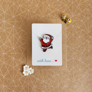 May include: A white card with a red and white cartoon Santa Claus enamel pin. The card has the text "with love..." written in blue with a red heart.