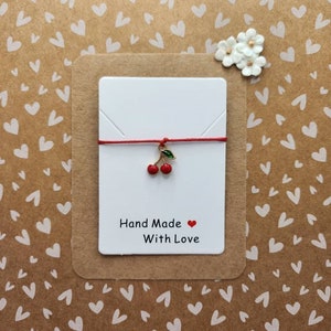 Pode incluir: Pulseira de cordão vermelho com um charme de cereja dourado e um cartão branco que diz "Hand Made With Love".