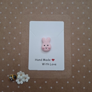 Puede incluir: Un dije en forma de conejito rosa sobre una tarjeta blanca con el texto "Hand Made With Love". La tarjeta está sobre un fondo marrón con lunares blancos. También hay una pequeña abeja y flores blancas.