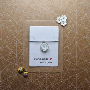 Puede incluir: Una tarjeta blanca con un dije de reloj plateado en una cuerda. La tarjeta dice "Hand Made With Love" y tiene un corazón rojo.