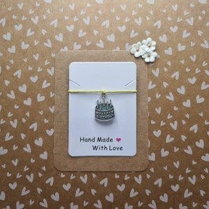 Pode incluir: Um cordão amarelo com um amuleto de prata de um bolo com as palavras "Happy Birthday" nele. O cordão está preso a um cartão branco com as palavras "Hand Made with Love" nele.