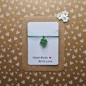 Może przedstawiać: Zielony czterolistny koniczyny urok na zielonym sznurku z białą kartką, na której napisano "Hand Made With Love" na brązowym tle z białymi sercami.