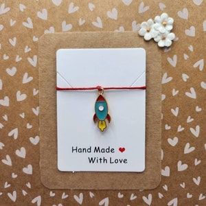 May include: A red string bracelet with a small gold rocket charm. The charm is blue, yellow, and red. The bracelet is on a white card with the text "Hand Made With Love" and a red heart.