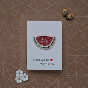 Puede incluir: Un broche de esmalte con forma de rodaja de sandía con detalles rojos, verdes y negros, expuesto en una tarjeta blanca. La tarjeta dice "Hand Made With Love". También hay dos pendientes con forma de abeja y adornos florales blancos.
