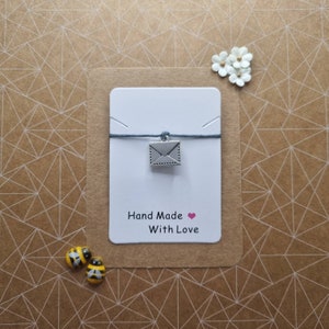 Pode incluir: Um bracelete de corda cinza com um amuleto de envelope prateado. O cartão atrás do bracelete diz "Hand Made with Love".