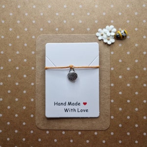 May include: A white card with the text "Hand Made With Love" and a red heart. A small silver seashell charm hangs from a thin orange string on the card.