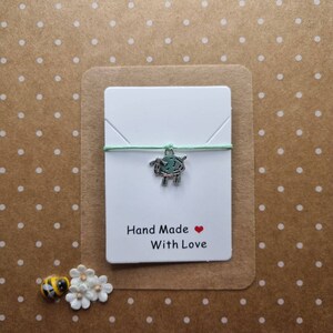 Puede incluir: Una tarjeta blanca con una cuerda verde y un dije de plata de una oveja. La tarjeta dice "Hand Made With Love" con un corazón rojo.
