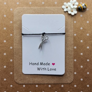 Può includere: Un piccolo ciondolo a forma di forbici argentate è appeso a un cordino nero, presentato su una carta bianca con la scritta "Hand Made With Love". La carta è posta su uno sfondo marrone a pois bianchi, con fiori decorativi e un'ape.