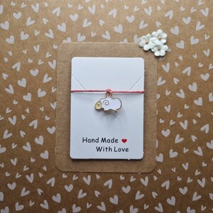 May include: A white card with a pink string and a gold sheep charm. The card says "Hand Made with Love" and has a red heart.