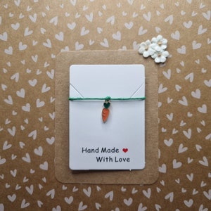 Peut inclure: Un bracelet en cordon vert avec un charme de carotte en or. Le bracelet est sur une carte blanche avec le texte "Hand Made With Love" et un cœur rouge.