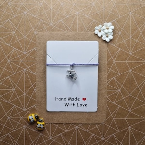 May include: A handmade witch-themed bracelet featuring a silver charm of a witch on a broomstick, strung on a purple cord. The bracelet is displayed on a white card with the text "Hand Made With Love" and a red heart. Two bee-shaped beads are at the bottom.