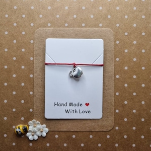 Pode incluir: Um bracelete de cordão vermelho com um amuleto de maçã de prata. O cartão diz "Hand Made With Love" e tem um coração vermelho.