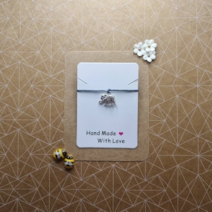 Puede incluir: Una tarjeta blanca con el texto "Hand Made With Love" y un corazón rosa. Una cuerda gris con un dije de elefante plateado está unida a la tarjeta.