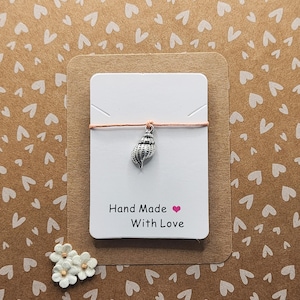 Peut inclure: Une carte blanche avec le texte "Hand Made With Love" et un cœur rose. Un charme en forme de coquillage argenté pend à une fine ficelle rose.