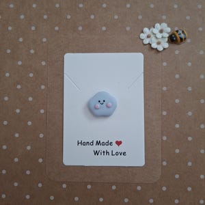 Pode incluir: Um cartão feito à mão com um enfeite em forma de nuvem azul claro com um rosto sorridente. O cartão diz "Hand Made With Love". O fundo é uma superfície castanha com bolinhas brancas.
