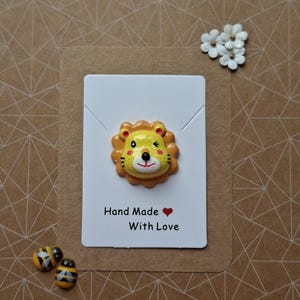 May include: A yellow and orange lion-shaped pin attached to a white rectangular display card. The card features the black text "Hand Made" followed by a red heart symbol and the text "With Love" printed in a simple sans-serif font.