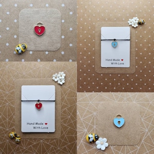 May include: Four images showing a red and blue heart-shaped lock charm on a black string bracelet. Each bracelet is displayed on a white card with the text "Hand Made With Love".