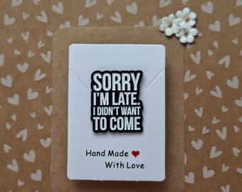 May include: A black and white enamel pin featuring the text SORRY I'M LATE. I DIDN'T WANT TO COME. The pin is attached to a white rectangular display card that reads Hand Made with a red heart symbol and the words With Love at the bottom.