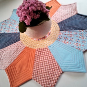 Könnte beinhalten: Ein farbenfroher, gesteppter Tischläufer mit einem Sternmuster. Der Läufer besteht aus verschiedenen Stoffen in Blautönen, Rosa, Orange und Gelb. Die Mitte des Läufers zeigt ein kreisförmiges Design mit mehreren Stoffschichten. Eine kleine Topfpflanze steht auf dem Läufer.